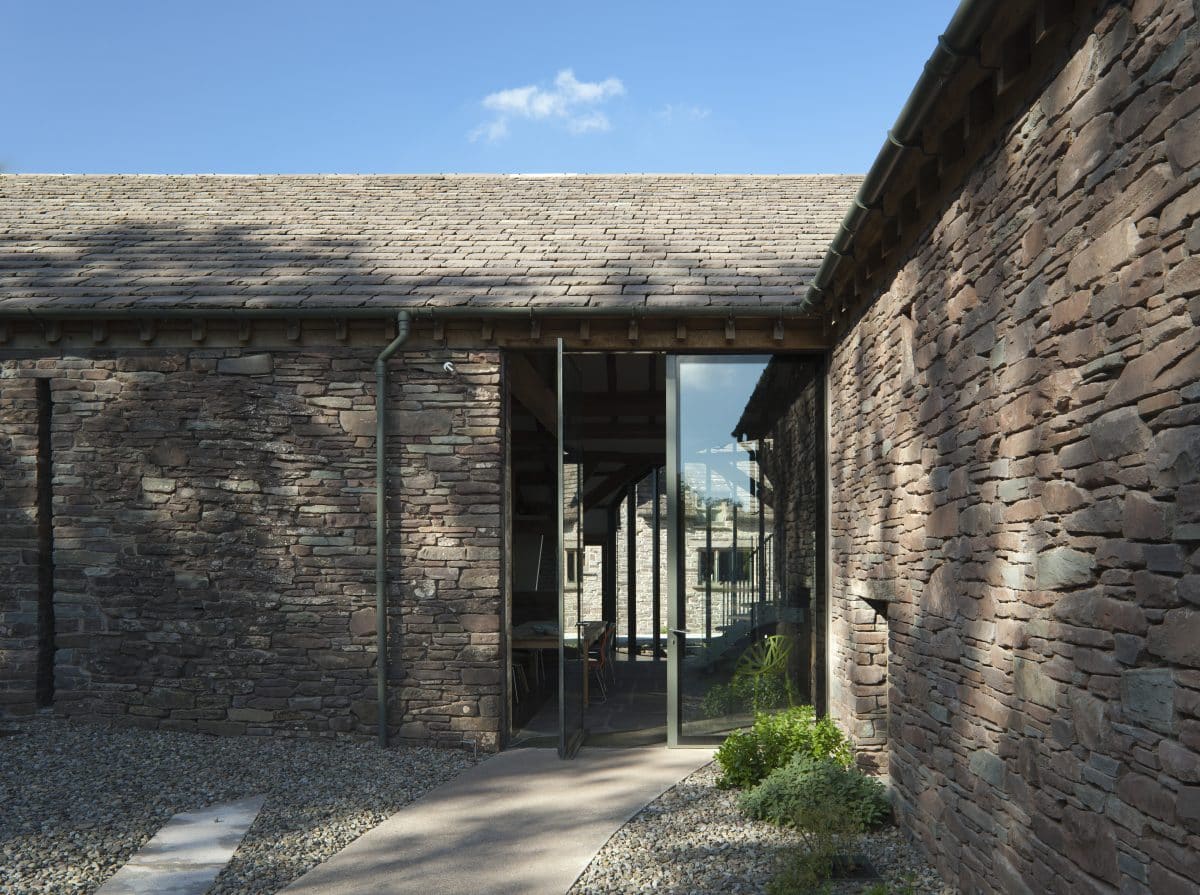 Barn Conversion. Perpendicular corner of L-shaped barn. The original stone walls have stood for an age and there is a two story window where the two walls join. The slate roof catches the sun in the car blue sky. Communion Architects, Hereford.
