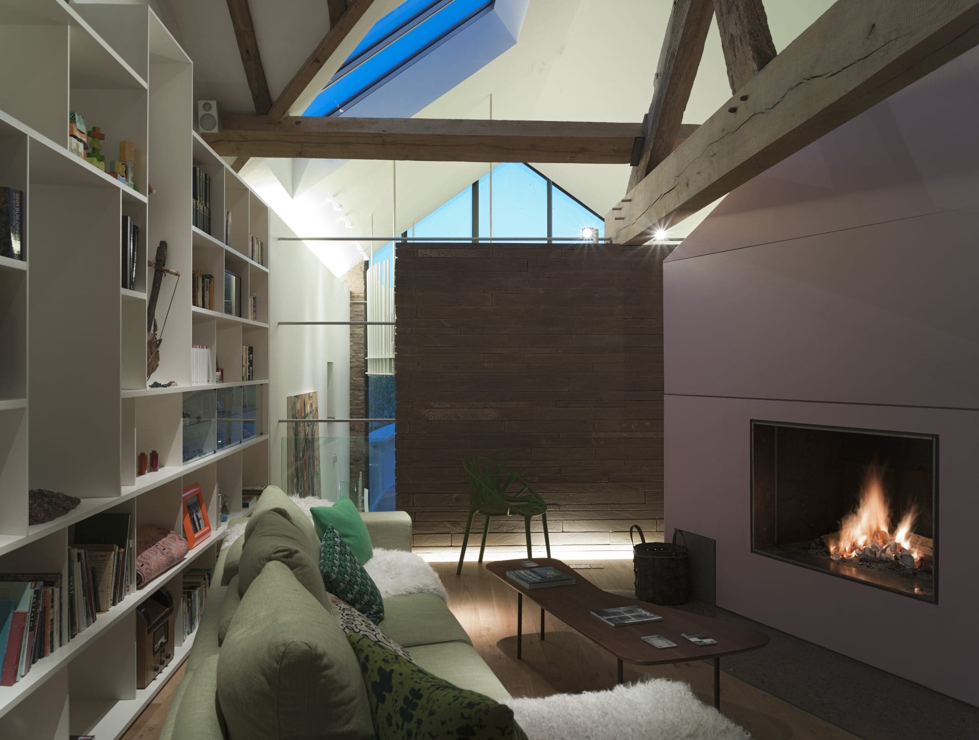 Interior of barn conversion. Left hand sided is an open bookcase, in front is a green sofa, which sits in front of am open fire, set into a lilac wall on the right hand side of the frame.