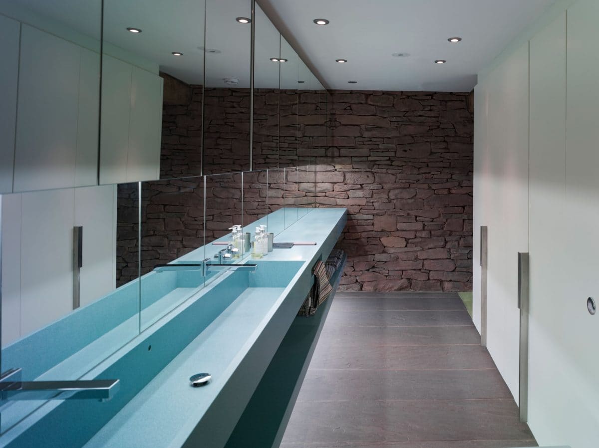 Looking along a bathroom in a barn conversion. The Left hand wall is covered in mirror from the ceiling down to the long blue steel sink with two silver taps. The end wall is original stone from the 15th barn. The right hand side was pale blue floor to ceiling doors for the cubicles. Barn conversion, communion architects Herefordshire 