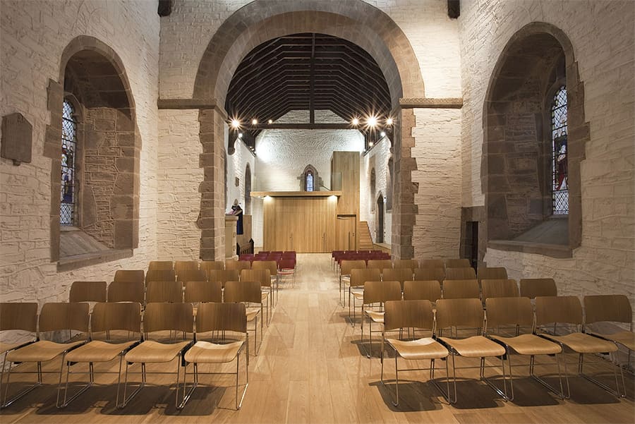 church re-ordering interior, looking down the central nave. Walls painted white with exposed stone framing the windows and central arch. A wooden flooring supports a rows of chairs, with space left for the central aisle. At the far end of the nave is a wood cube-like structure, this contains a kitchen area (unseen).