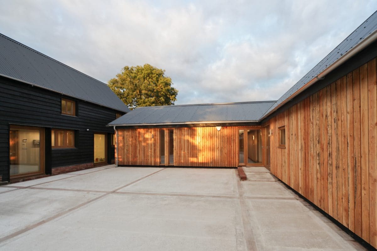 Perpendicular corner of an L-shaped barn conversion. The single storey exterior is rendered with vertical planks of timber, interrupted with full height glazing. The warm evening light creates a warm, orange image for this barn conversion and business, Communion Architects, Herefordshire.