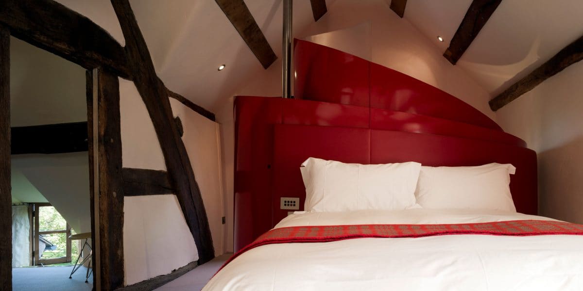 A white bed in the centre of the frame with a curving red steel structure behind, behind this is a bathroom unseen, the room is painted white with exposed timber beams. 15th century farm house, barn conversions.