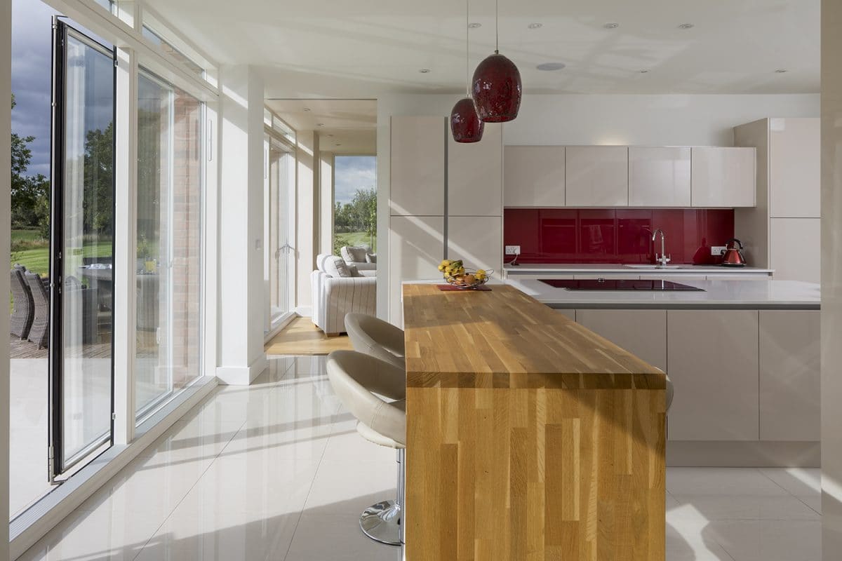 Interior of a home extension, left hand wall open bookcase, right hand wall and far back wall is glazed with timber frames, in the centre are two grey sofas on a oak floor.