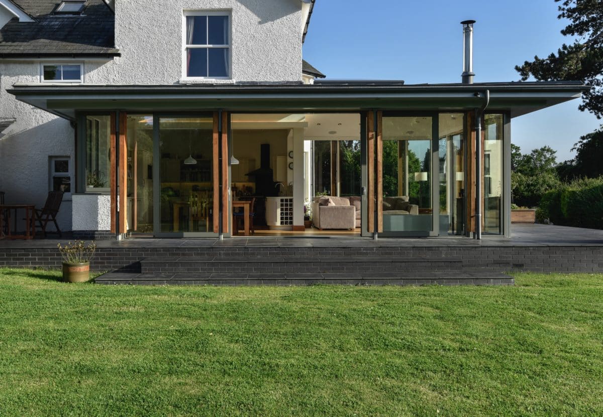 Tripartite image of grass, glazed extension and the original house and sky in the upper third. The doors to the glazed and aluminium extension are open, welcoming people in. Communion Architects, Herefordshire.