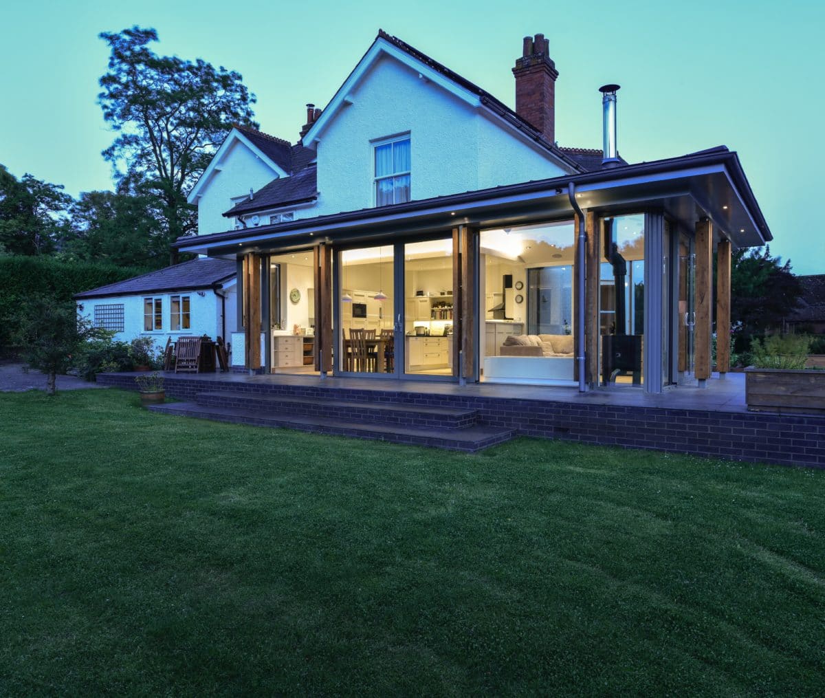 Exterior of home extension at twilight. The warm lighting inside the glazed extension brings focus to it, with the family dining room table inside, the original, white 1930s house rises behind. Communion Architects, Hereford.