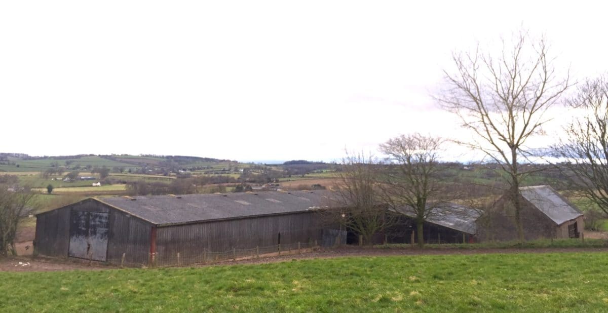 At the bottom of a green field is a long dark grey barn. Industrial style agricultural barn with potential for a barn conversion. The view behind shows the beautiful rolling hills of Herefordshire. transforming space, Communion Architects, Herefordshire. 