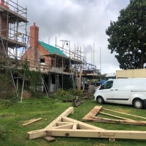 A wooden oak roof truss lays on green grass in the foreground, behind it is a white van and a red brick cottage covered with scaffolding. Communion Architects, Hereford.