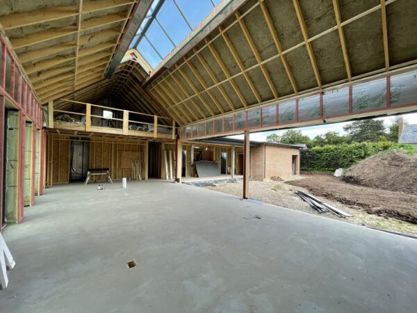 Interior of a new dwelling in Herefordshire, under construction. Concrete floor, timber beams, exposed insulation, in the pitched roof. The pitched roof has a pitched window running the length of the peak. The walls show exposed breeze blocks and brick, with the right hand wall open for floor to ceiling windows. Communion Architects Hereford.