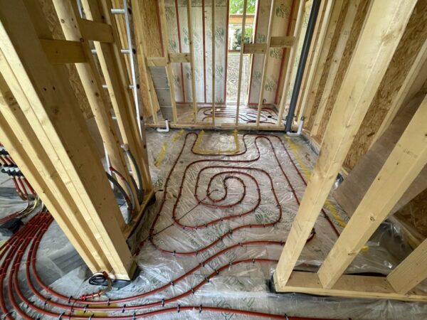 Timber framed wall building, part way through construction, with no packing between the timber frames yet. On the floor is a curling red tube curling across the floor expanse - part of an underfloor heating system. communion Architects Hereford, Herefordshire.