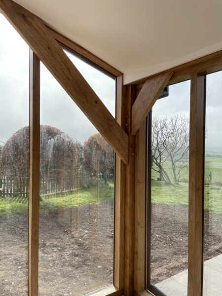 A portrait image of a corner of a room where two floor to ceiling windows meet with oak beam framing and bracing the space. cream roof and garden outside with a miuddy flower bed, grass and trees beyond. Communion Architects, Hereford, Herefordshire.