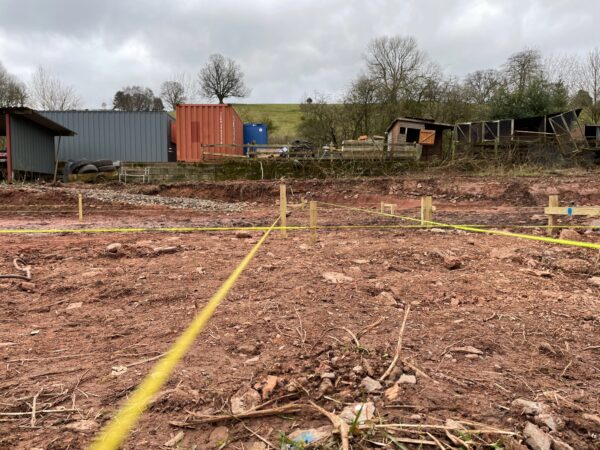 A muddy field, red Herefordshire clay soil, with yellow rope marking out rectangular zones. Preparing to dig foundations. Communion Architects, Hereford, Herefordshire.