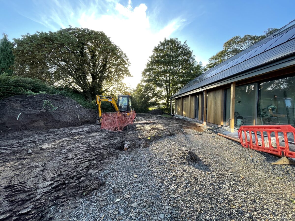 A small yellow digger clearing a muddy area while in the right hand side of the image is a new single storey house with a slate roof and glass and timber walls. Communion Architects Hereford.