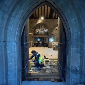 Church archway with the door open showing the inside of the church where a workman is kneeling, laying a new timber floor in the space. Communion Architects Hereford.