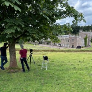 A photographer with camera on tripod, standing in beautiful countryside and waiting for a break in the clouds to get the perfect shot of a historic building. Communion Architects Hereford